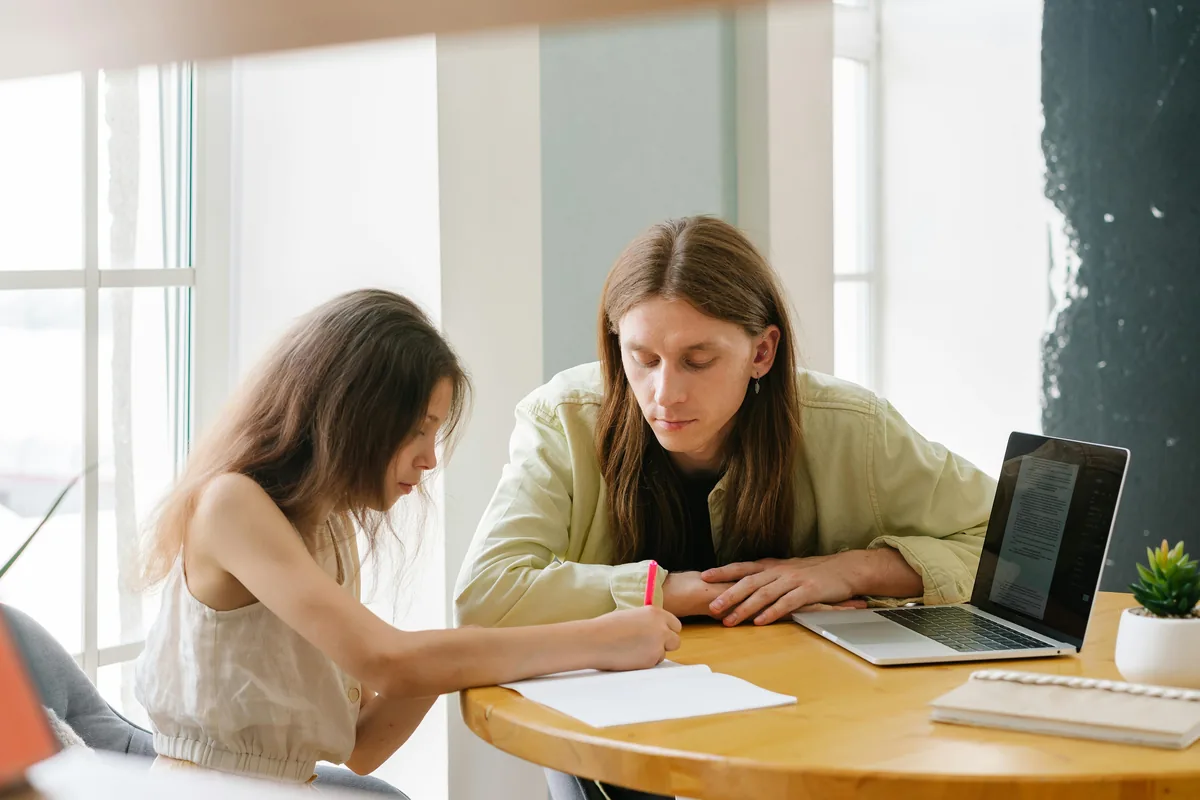 A parent sitting alongside their child at the dining table, an open laptop nearby and the child writing on paper — the everyday shape of free online homeschool programs in practice.