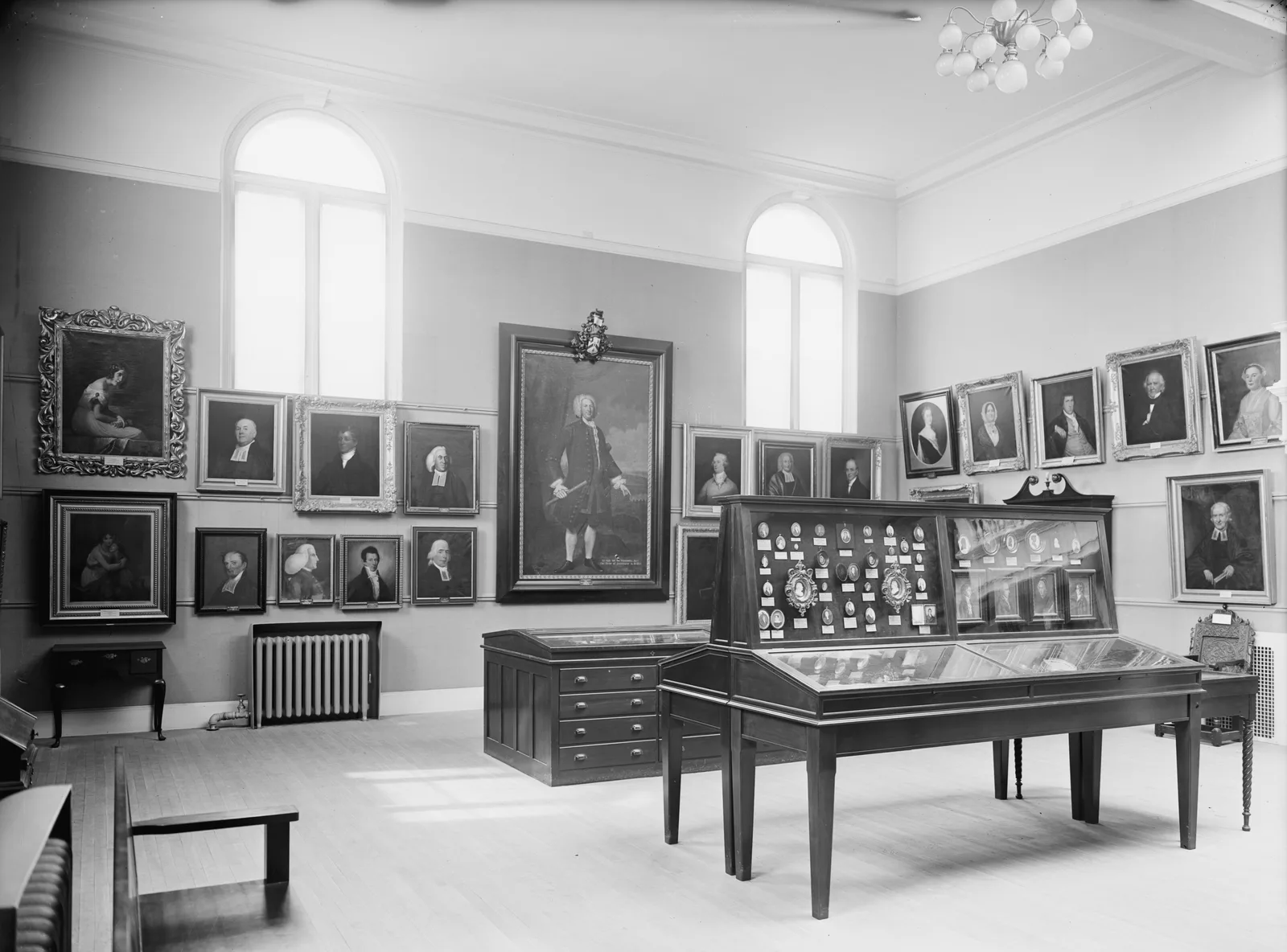 Black-and-white photograph of an empty portrait gallery: rows of framed oil portraits hung on the walls, tall arched windows, and a glazed case of portrait miniatures on a wooden stand