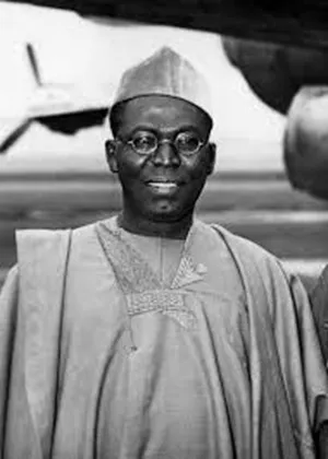 Black and white archive portrait of a bespectacled Nigerian leader in an embroidered agbada, photographed beside an aircraft