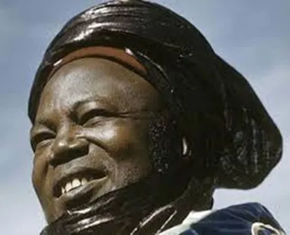 Archive portrait of a smiling Nigerian woman in a large dark gele headwrap, photographed against an open sky